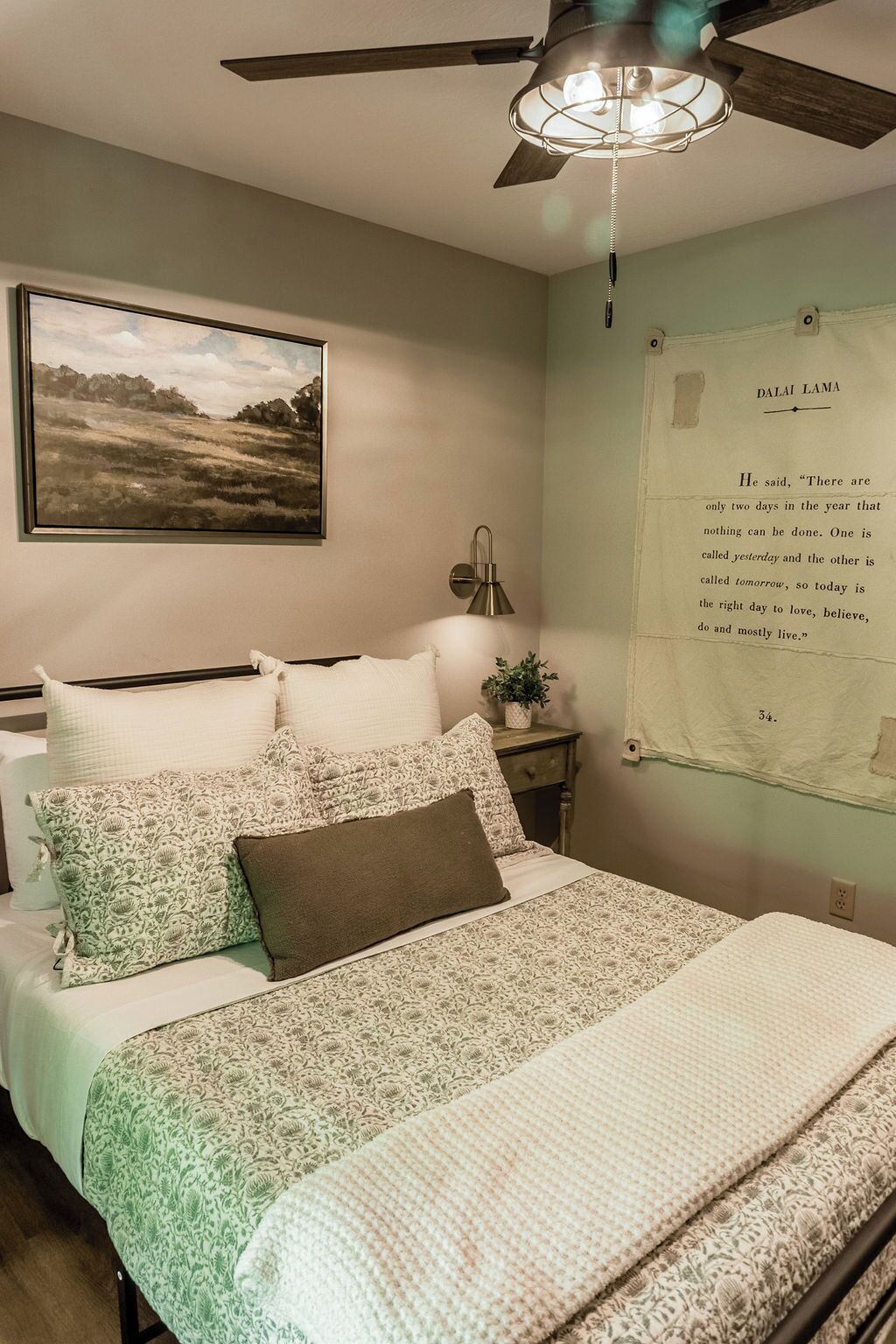 A bedroom with a bed, a ceiling fan, a wall-mounted lamp, a framed quote, and a painting.