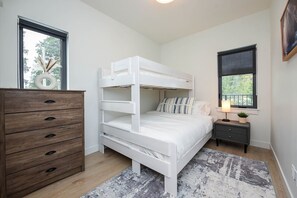 A bunk bed with a wooden dresser, a bedside table with a lamp, and a window with a view of trees.