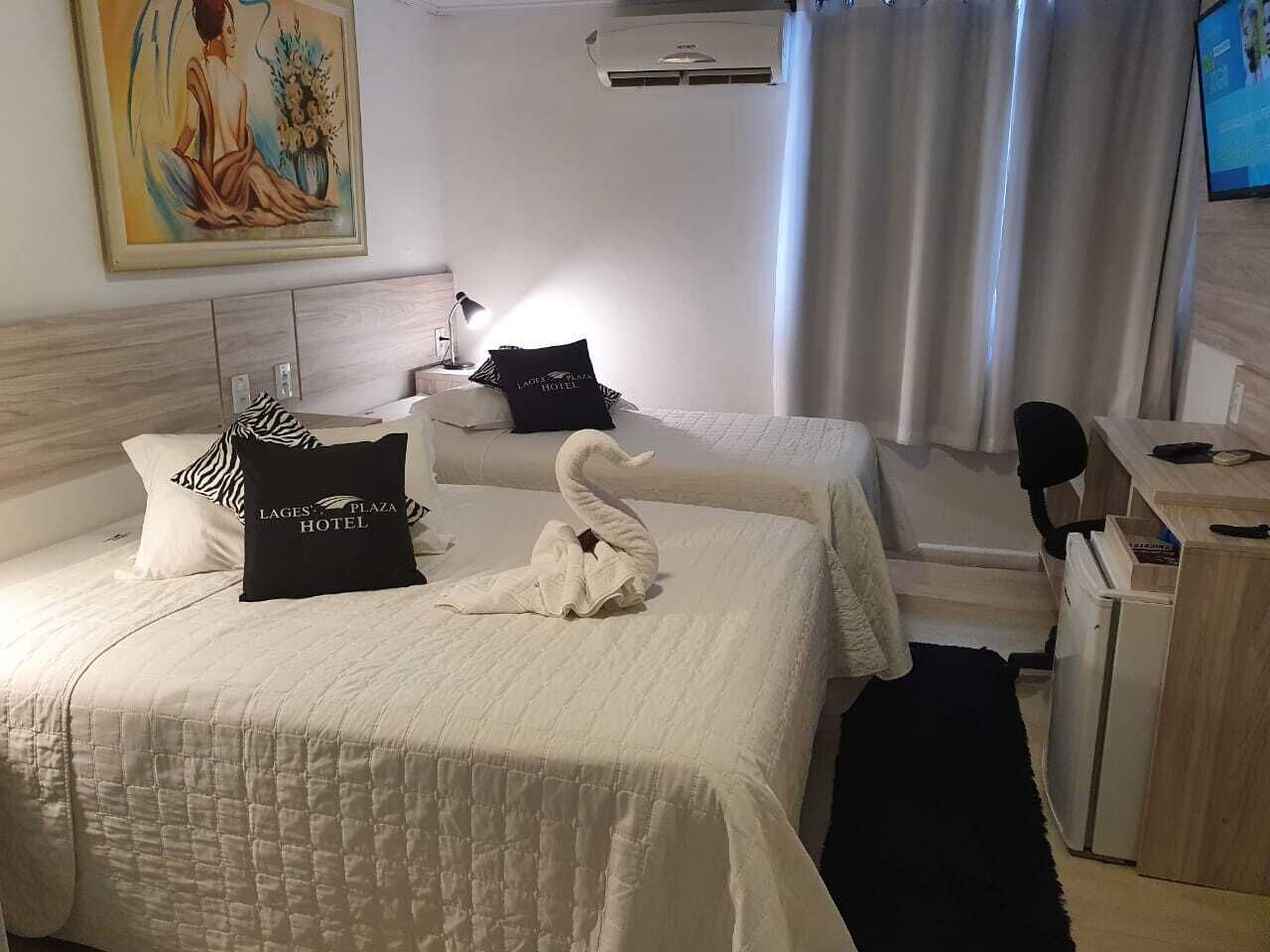 A hotel room with two beds, a desk, and a chair. There is a painting on the wall and an air conditioning unit above the bed.