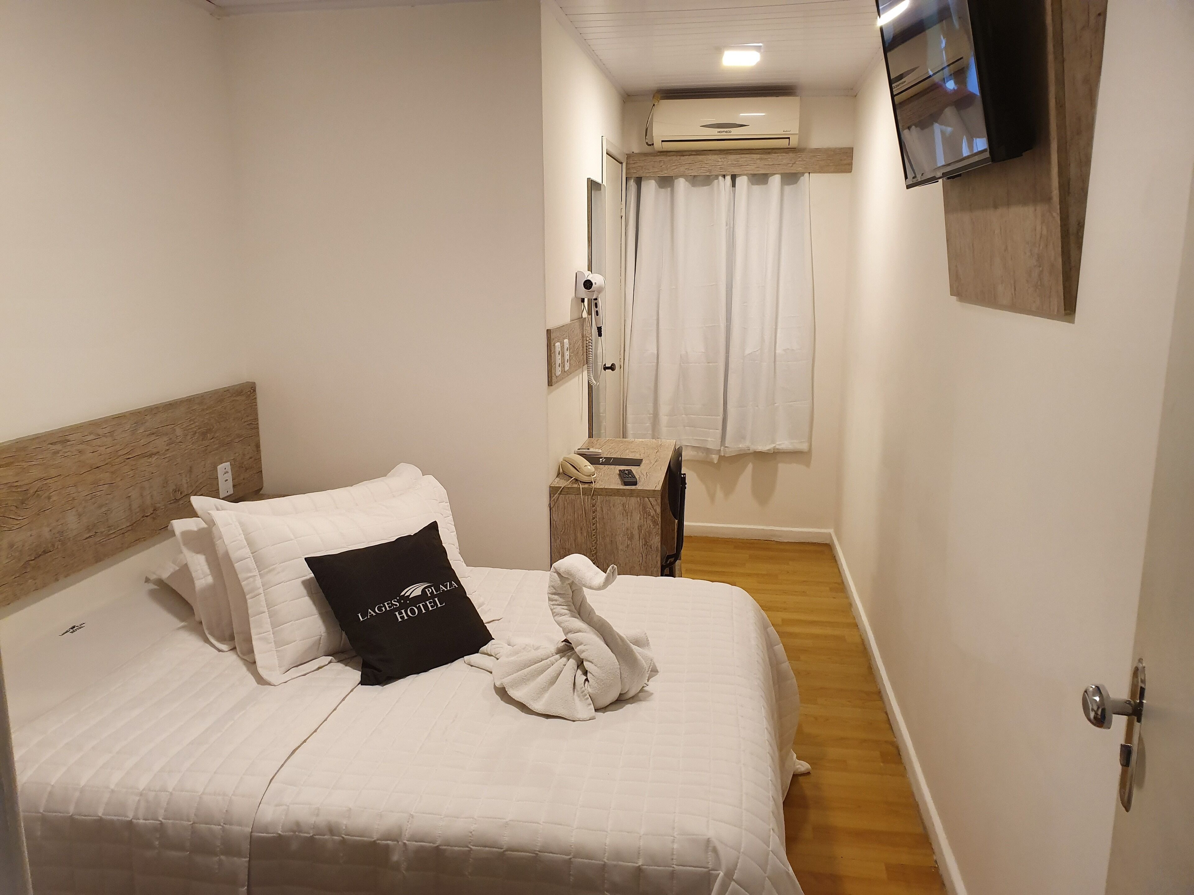 A hotel room with a bed, a wooden headboard, a pillow with the hotel's name, a towel, a bedside table with a phone and remote, a mirror, and a mounted TV.