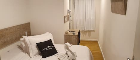 A hotel room with a bed, a wooden headboard, a pillow with the hotel's name, a towel, a bedside table with a phone and remote, a mirror, and a mounted TV.