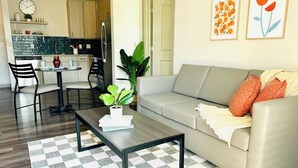 A modern living room with a sofa, coffee table, and chairs. There is a kitchen in the background with cabinets and a clock.