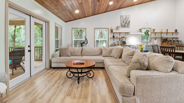 A spacious living room with a wooden ceiling, a large beige sectional sofa, a round coffee table, and a sliding glass door leading to an outdoor deck. - Sea Cow Retreat - Near Springs + Porch, Covered Deck & Fire Pit (Crystal River)