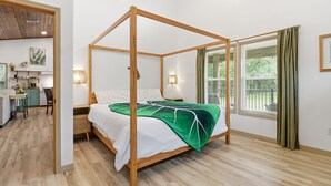A four-poster bed with a green leaf-patterned bedspread, wooden flooring, a kitchen area with a dining table, and a view of greenery through the window. - Sea Cow Retreat - Near Springs + Porch, Covered Deck & Fire Pit (Crystal River)