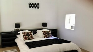 Quarto moderno com estrutura de cama preta, roupa de cama branca e almofadas estampadas. Há duas mesas de cabeceira com luminárias, uma obra de arte na parede preta e uma janela com vista.