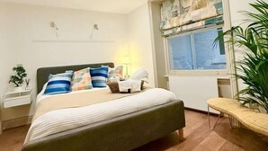 A modern bedroom with a large bed, a bedside table, a nightstand, a window with blinds, and a potted plant.
