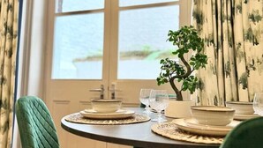 A dining area with a round table, green upholstered chairs, a bonsai tree, and a window with a view of a water body.