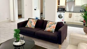 A modern living room with a dark brown sofa, a round glass coffee table, and a white sofa. A kitchen with white cabinets and a microwave is visible in the background.