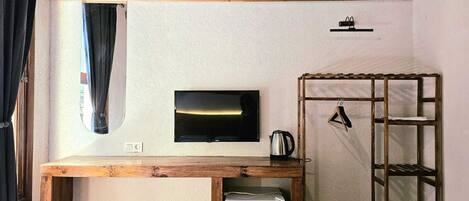 Una habitación compacta con un televisor montado en la pared, una pequeña mesa de madera con un hervidor y una estantería metálica.