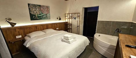 Quarto com cama, cabeceira de madeira, pintura, mesa de cabeceira, espelho e banheiro com banheira e vaso sanitário.
