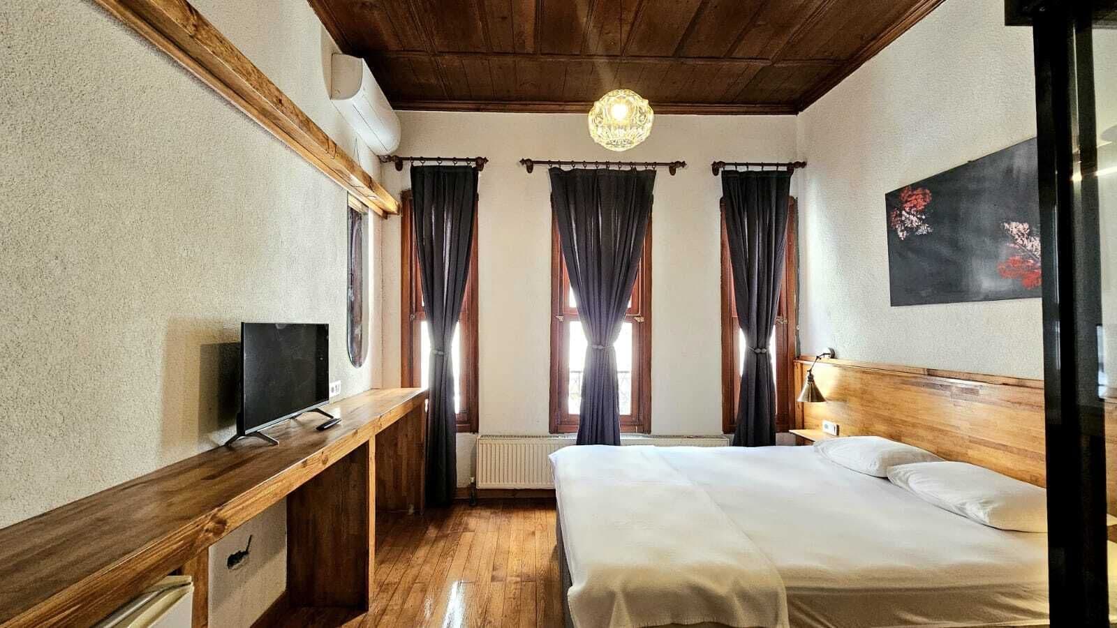 A hotel room with a wooden desk, a bed, a TV, and a window with curtains.