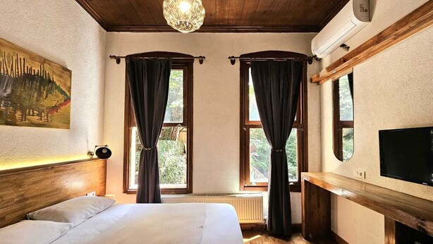 A bedroom with a bed, two windows with curtains, a painting, a ceiling light, and a TV on a wooden shelf. - Mahzen Otel (Istanbul)
