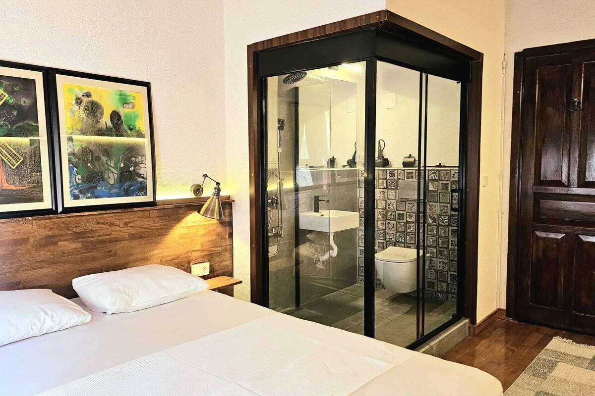 A hotel room with a bed, a wooden headboard, two framed artworks, a glass-enclosed bathroom, and a dark wooden door.