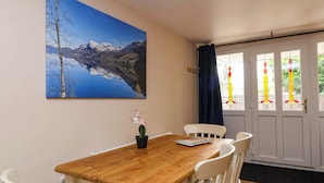 A dining area with a wooden table and white chairs, a painting of a mountain landscape, and a door with stained glass windows. - Poddington Cottage (Salisbury)