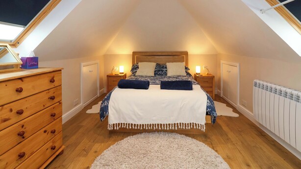 A bedroom with a sloped ceiling, a bed with a white and blue bedspread, two bedside tables with lamps, a wooden dresser, and a radiator. - Poddington Cottage (Salisbury)