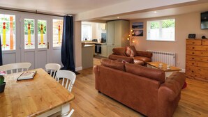 A cozy living room with a brown leather sofa, a wooden dining table with chairs, and a kitchen area in the background. - Poddington Cottage (Salisbury)