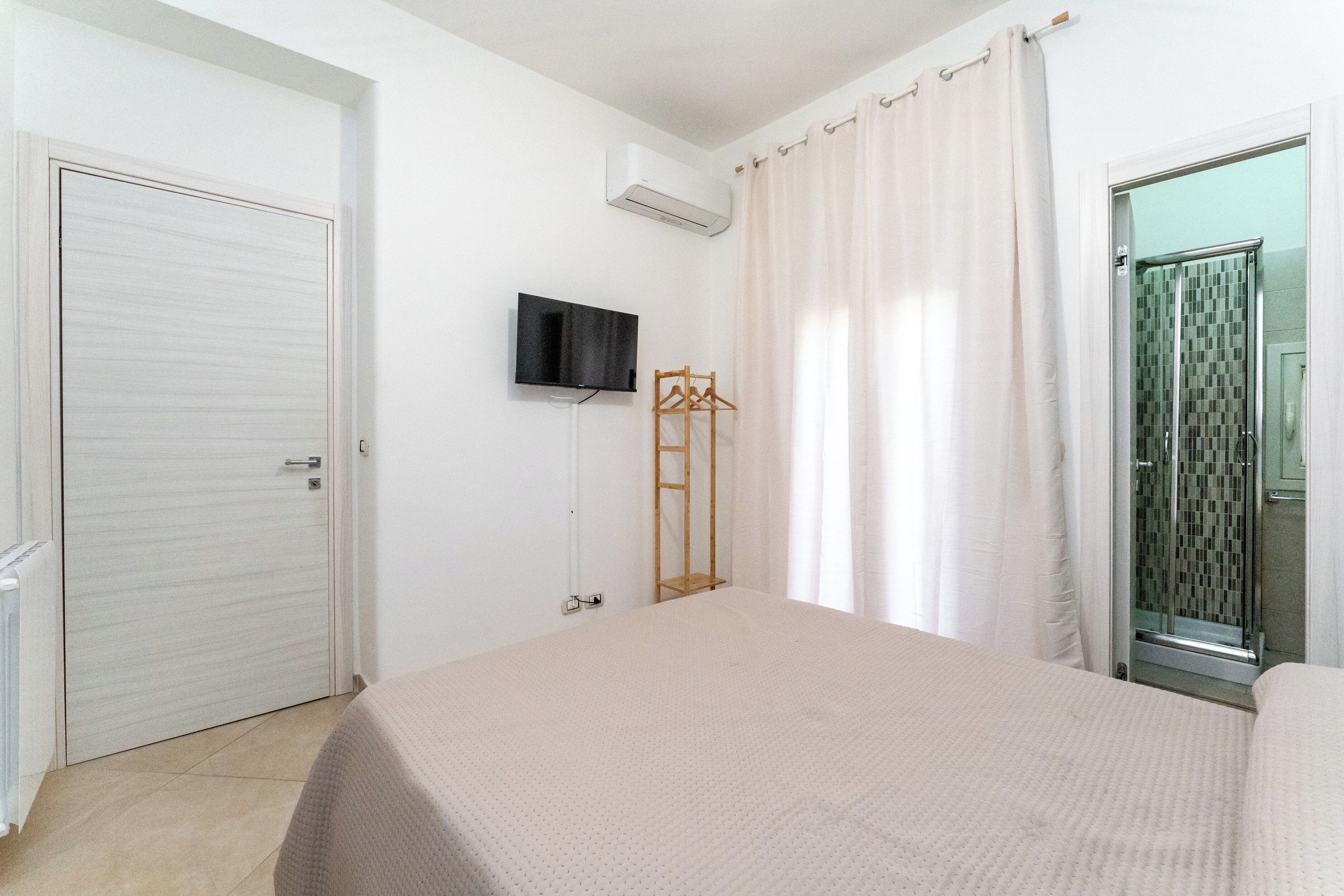 Een slaapkamer met een bed, een aan de muur bevestigde televisie, een houten kleerhanger en een badkamer met een douche.