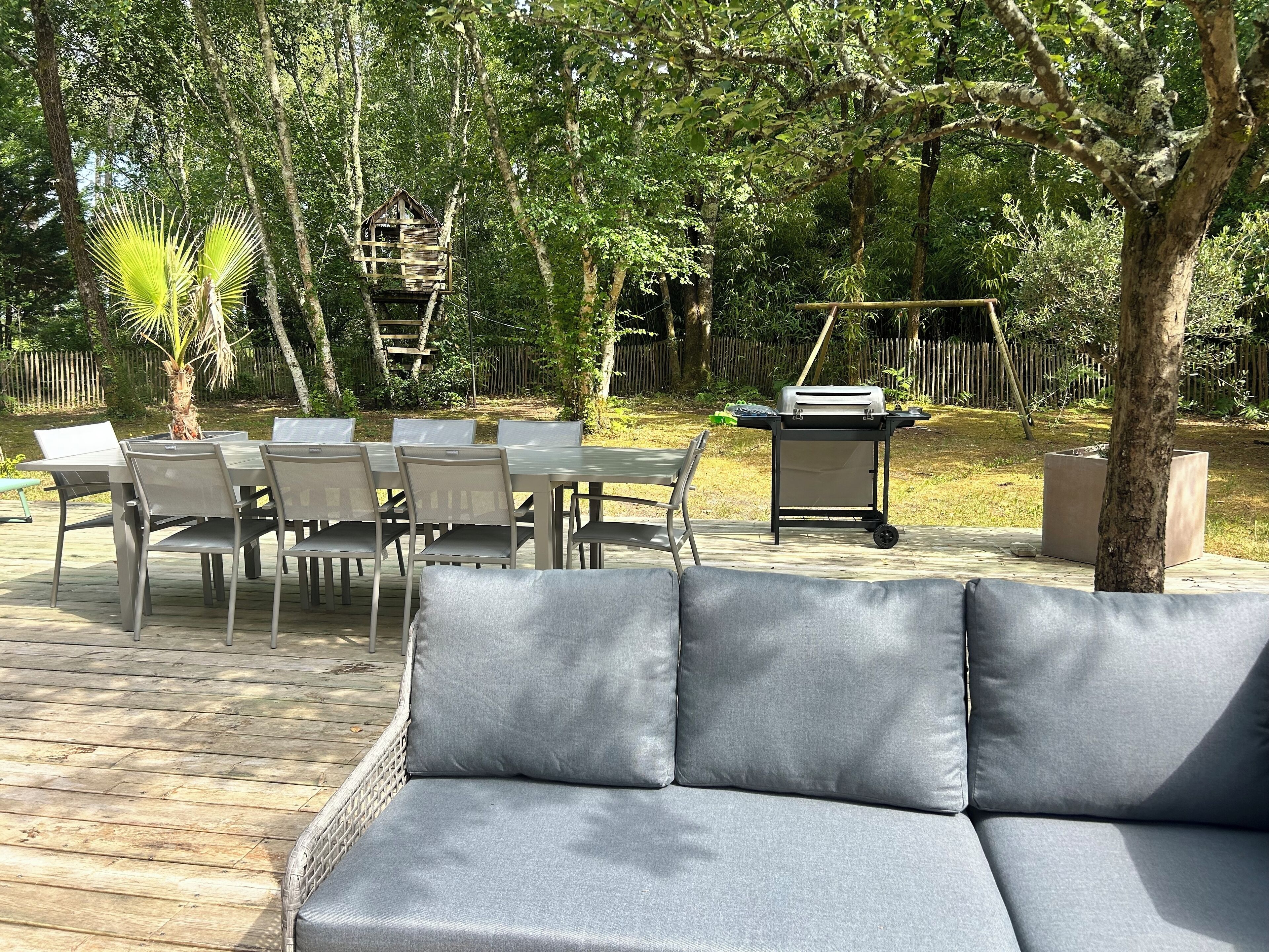 An outdoor patio with a grey sofa, a dining table with chairs, and a barbecue grill.