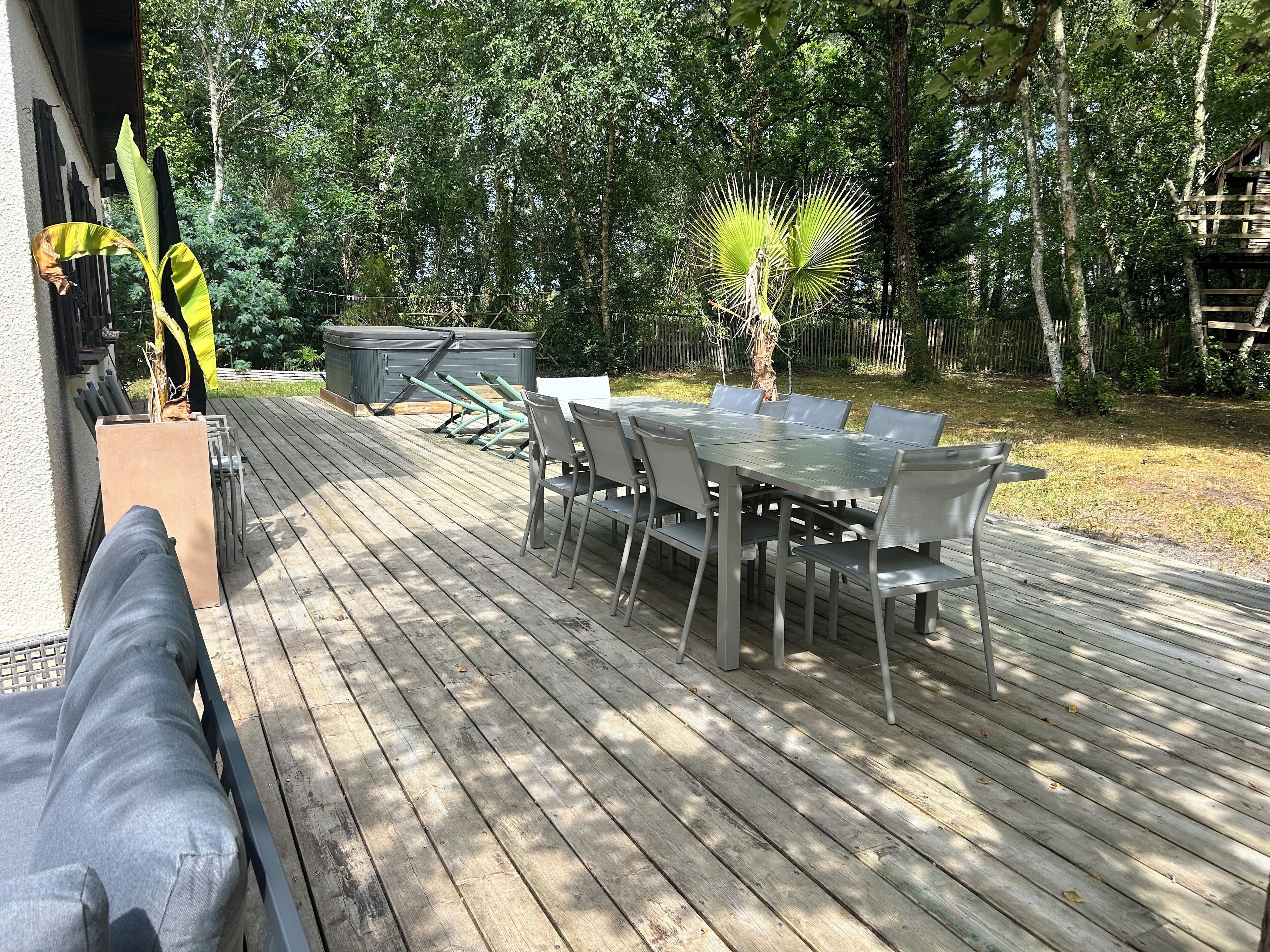 Um terraço ao ar livre com uma mesa de jantar e cadeiras, cercado por árvores e uma cerca de madeira.