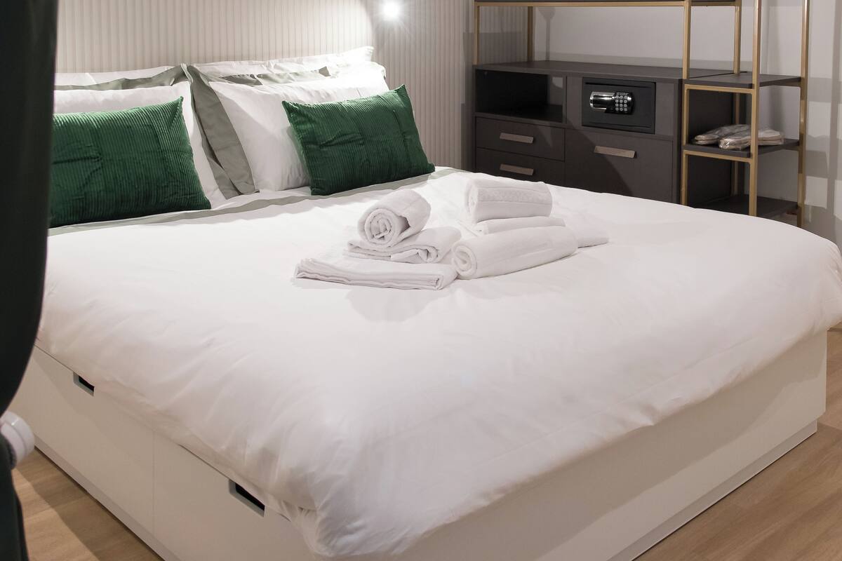 A neatly made bed with white linens and rolled towels, a bedside console with a lock, and a wooden nightstand with a mirror.