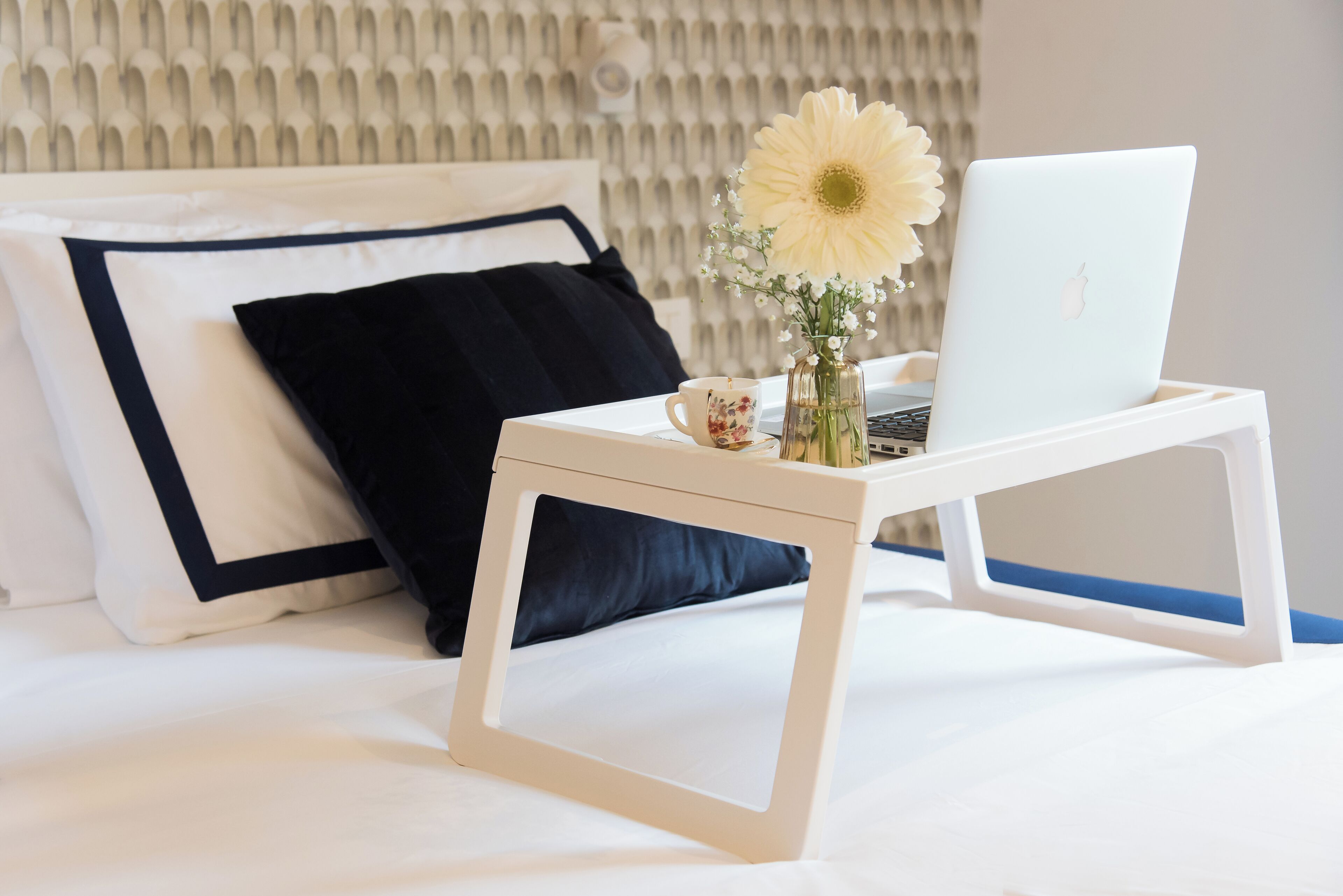 A neatly made bed with a laptop on a bedside table, a cup, and a vase with flowers.