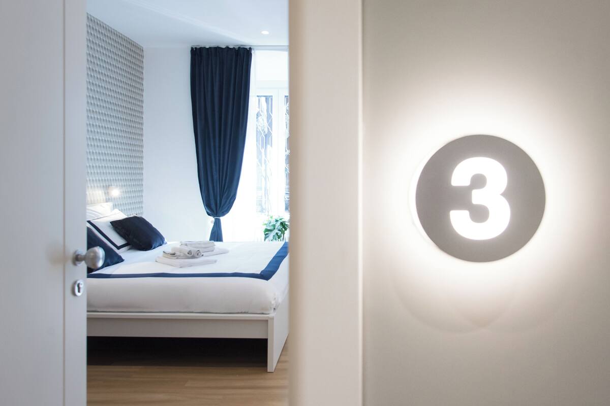 A hotel room with a bed, a window with curtains, and a wall-mounted number sign.