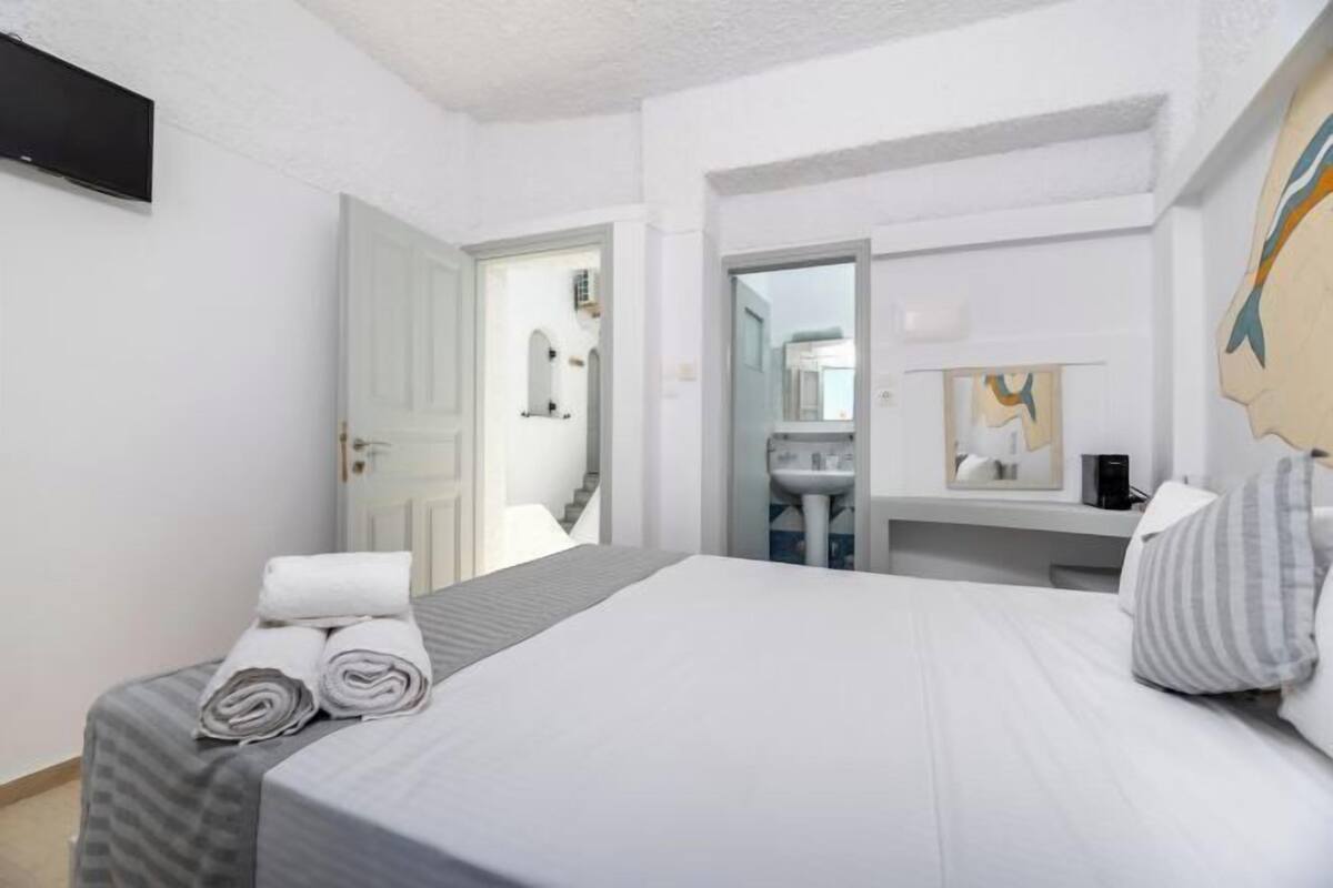 A hotel room with a bed, a desk with a lamp, a TV, and a bathroom visible through an open door.