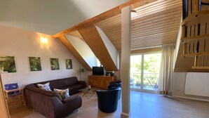 A spacious living area with a high ceiling, wooden beams, a brown sofa, a television, and a view to the outside.