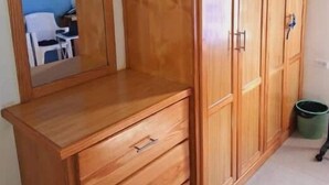 A wooden wardrobe with multiple doors and drawers, a mirror, and a green trash bin in a room with a blue wall and a white chair. - Breezy Bungalow Airport Suite (Piarco)