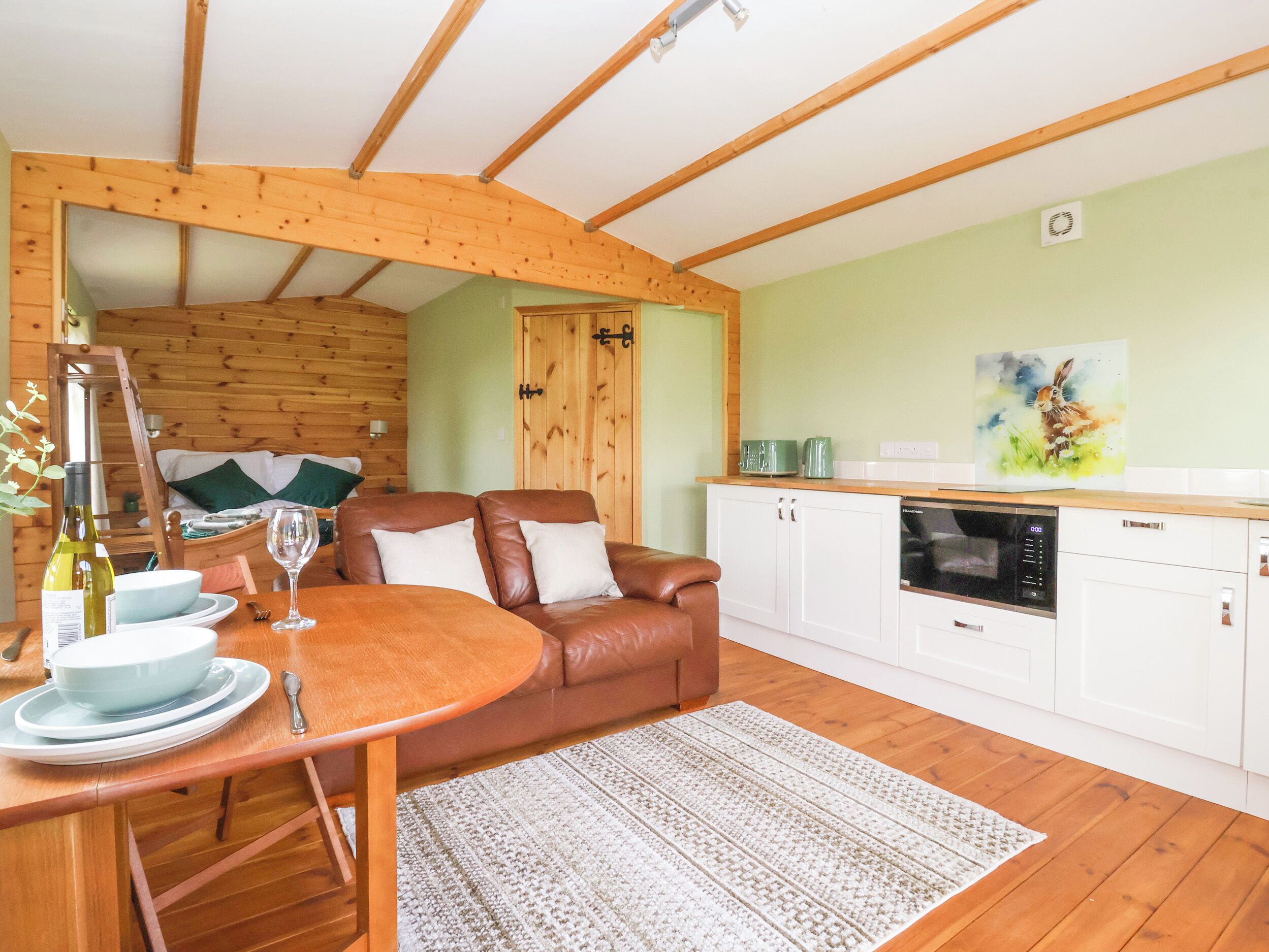 Una habitación acogedora con techo de madera, una mesa de comedor con sillas, un sofá de cuero marrón, una zona de cocina con armarios blancos y un cuadro en la pared.