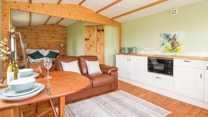 Een gezellige kamer met een houten plafond, een eettafel met stoelen, een bruine leren bank, een keuken met witte kasten, en een schilderij aan de muur.
