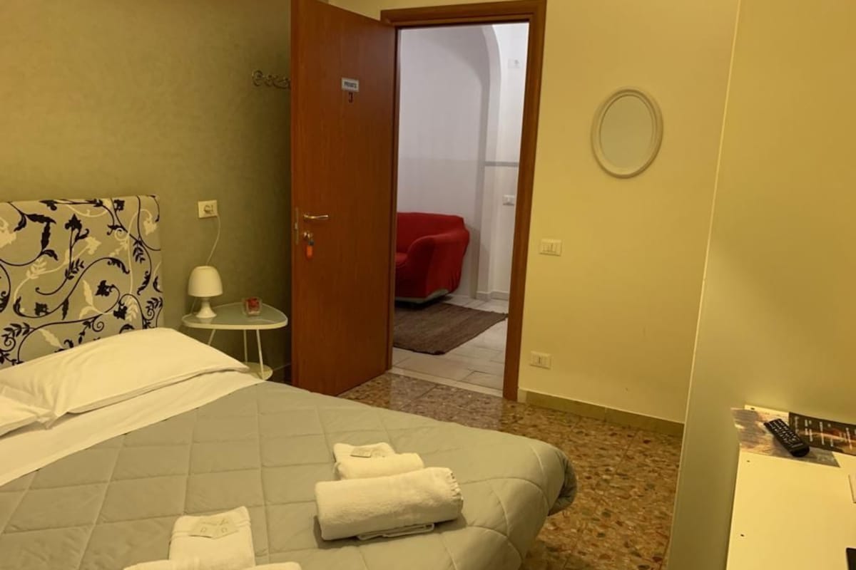 A hotel room with a bed, a bedside table with a lamp, a red chair in the adjacent room, and a round mirror on the wall.