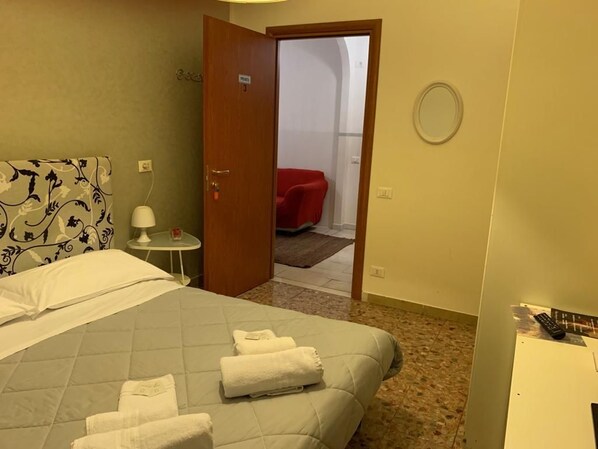 A hotel room with a bed, a bedside table with a lamp, a red chair in the adjacent room, and a round mirror on the wall. - Il Giardino di Gioia (Verona)