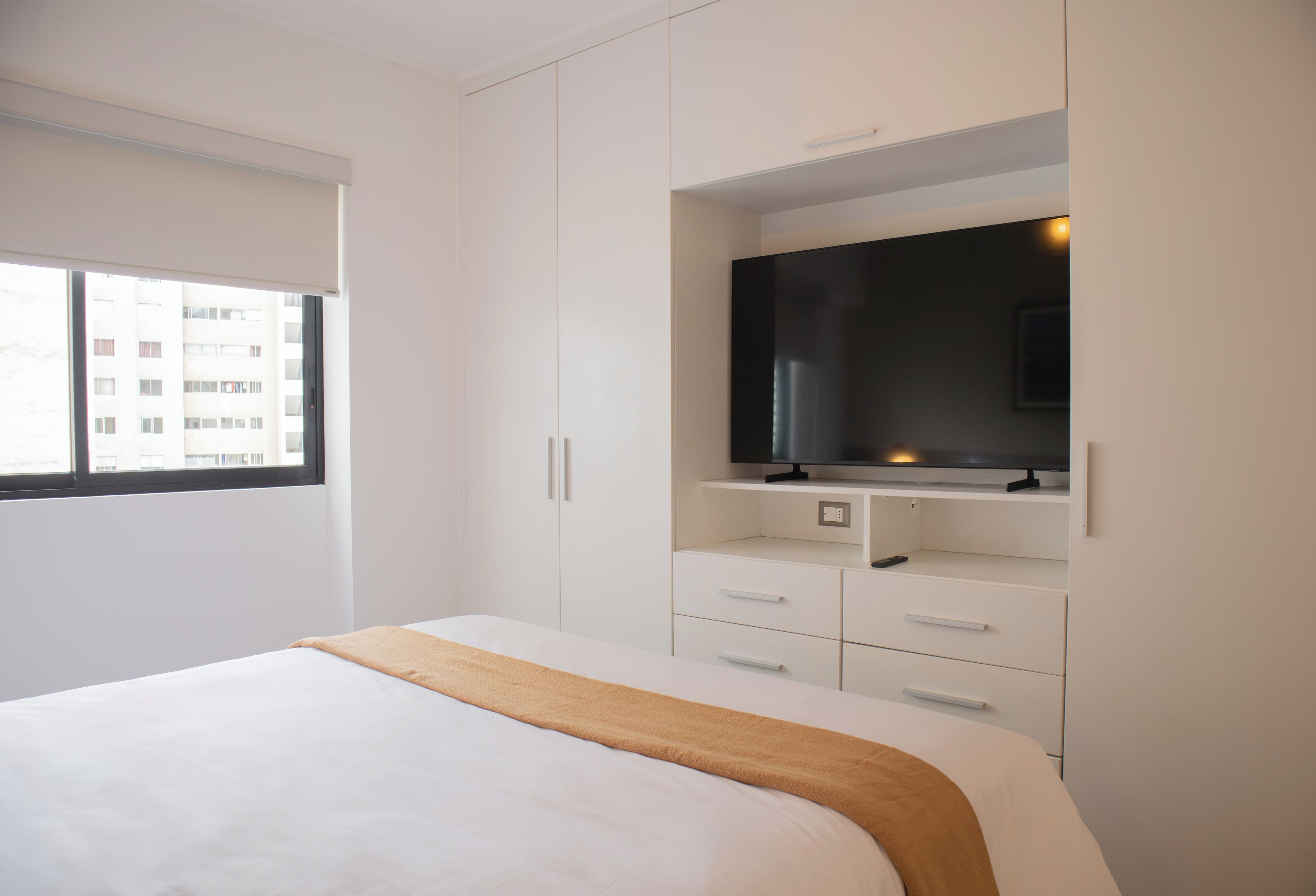 Un dormitorio con cama, un televisor montado en un mueble y una ventana con vistas a edificios.