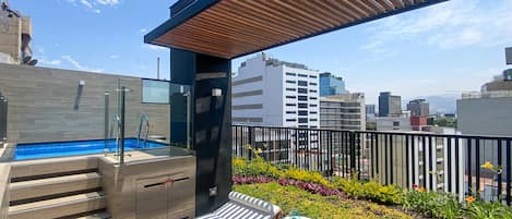 Un área de piscina en la azotea con cerramiento de vidrio, terraza de madera y sillas de descanso.