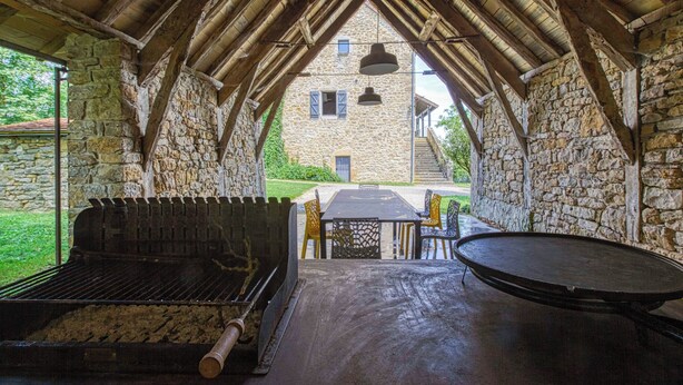 An outdoor patio with a stone wall, a grill, a round table, and a view of a stone building in the background. - Gite with swimming pool le Bervic (Sainte-Croix)