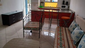 Une cuisine compacte équipée d’un réfrigérateur, d’un four à micro-ondes et d’un évier. On y trouve également un coin repas avec une table et des chaises, ainsi qu’un canapé agrémenté de coussins à motifs.