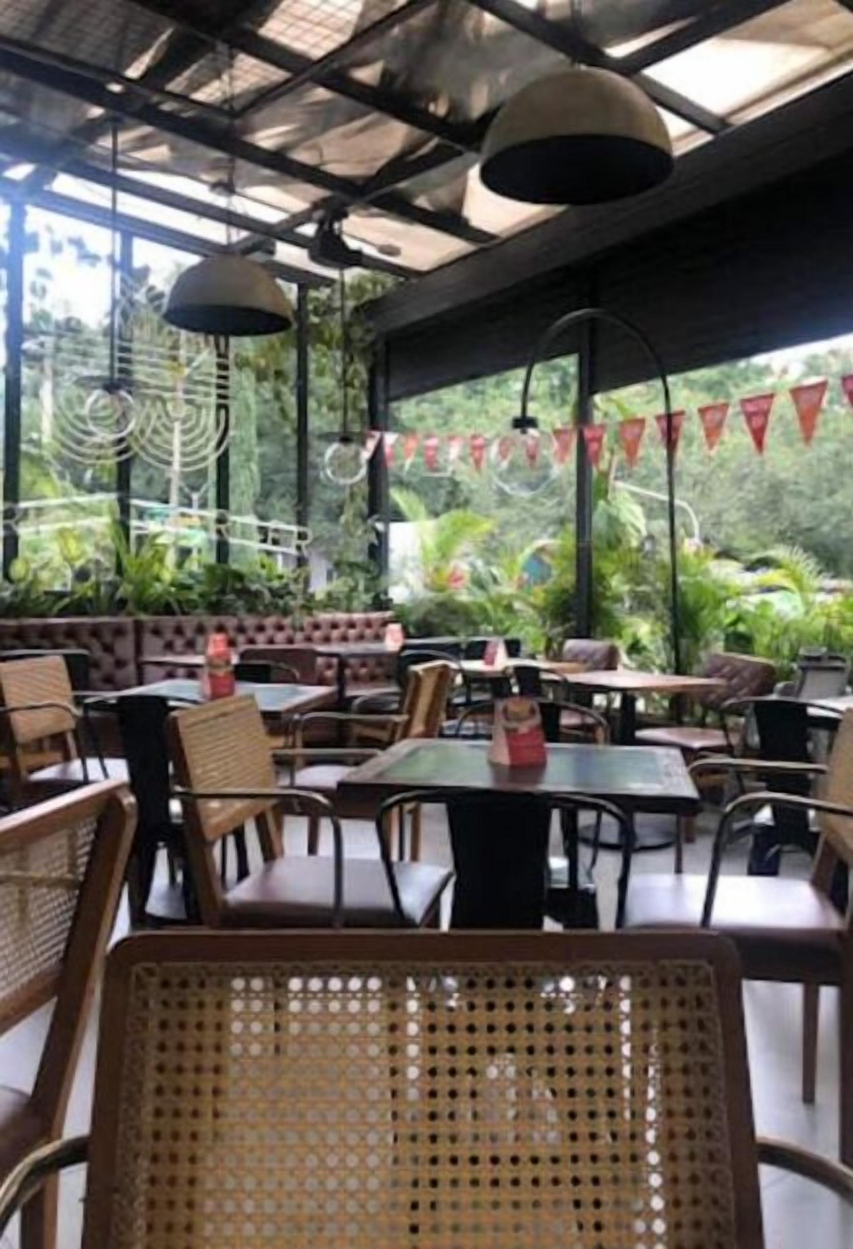 An outdoor cafe with tables and chairs, surrounded by greenery and a covered area with a metal roof.