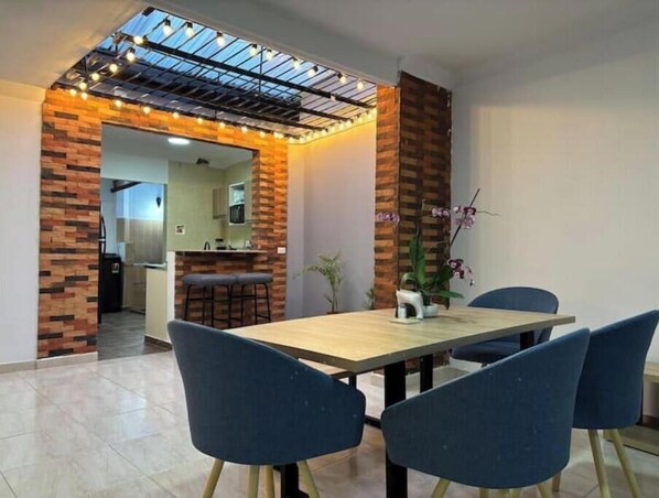 A dining area with a wooden table and blue chairs, a brick accent wall, and a kitchen in the background.
