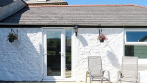 Un bâtiment à l’extérieur blanc, avec un toit recouvert de tuiles et une porte en verre. Il y a deux chaises à l’extérieur.