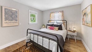 A bedroom with a bed, a nightstand, a window with blinds, a framed artwork on the wall, and a patterned rug.