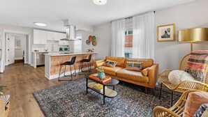 A modern living room with a sofa, coffee table, and wicker chairs. There is a kitchen area with white cabinets and a dining table.