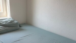A small, single-bed room with a plain white wall and a window on one side.
