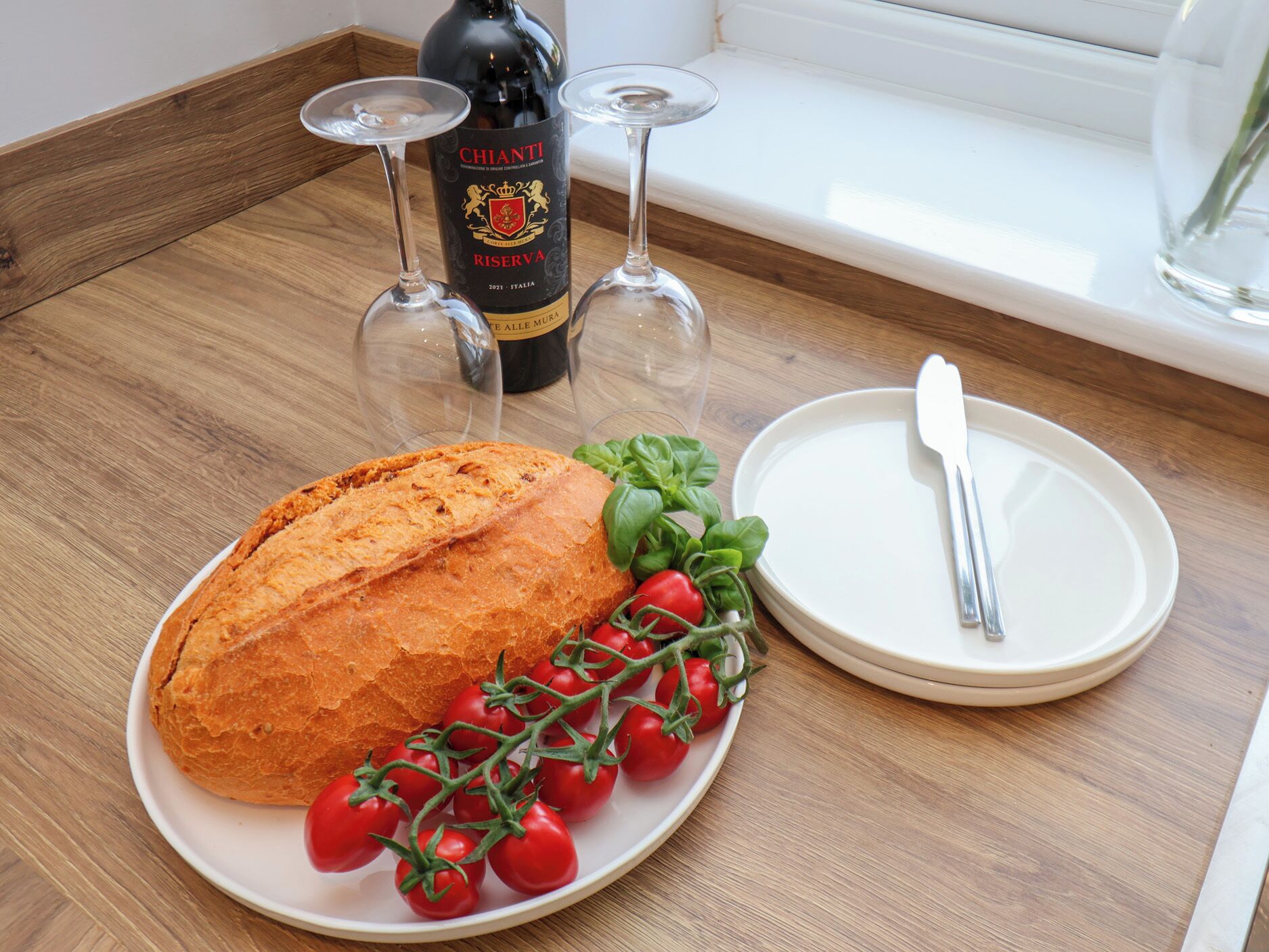 A loaf of bread on a plate with cherry tomatoes and basil, a bottle of Chianti wine, and two wine glasses on a wooden table.