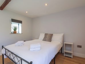 Ein ordentlich bezogenes Bett mit weißen Leinentüchern und gefalteten Handtüchern, ein Nachttisch, ein kleines Regal und ein Fenster mit Jalousien und einer Pflanze.