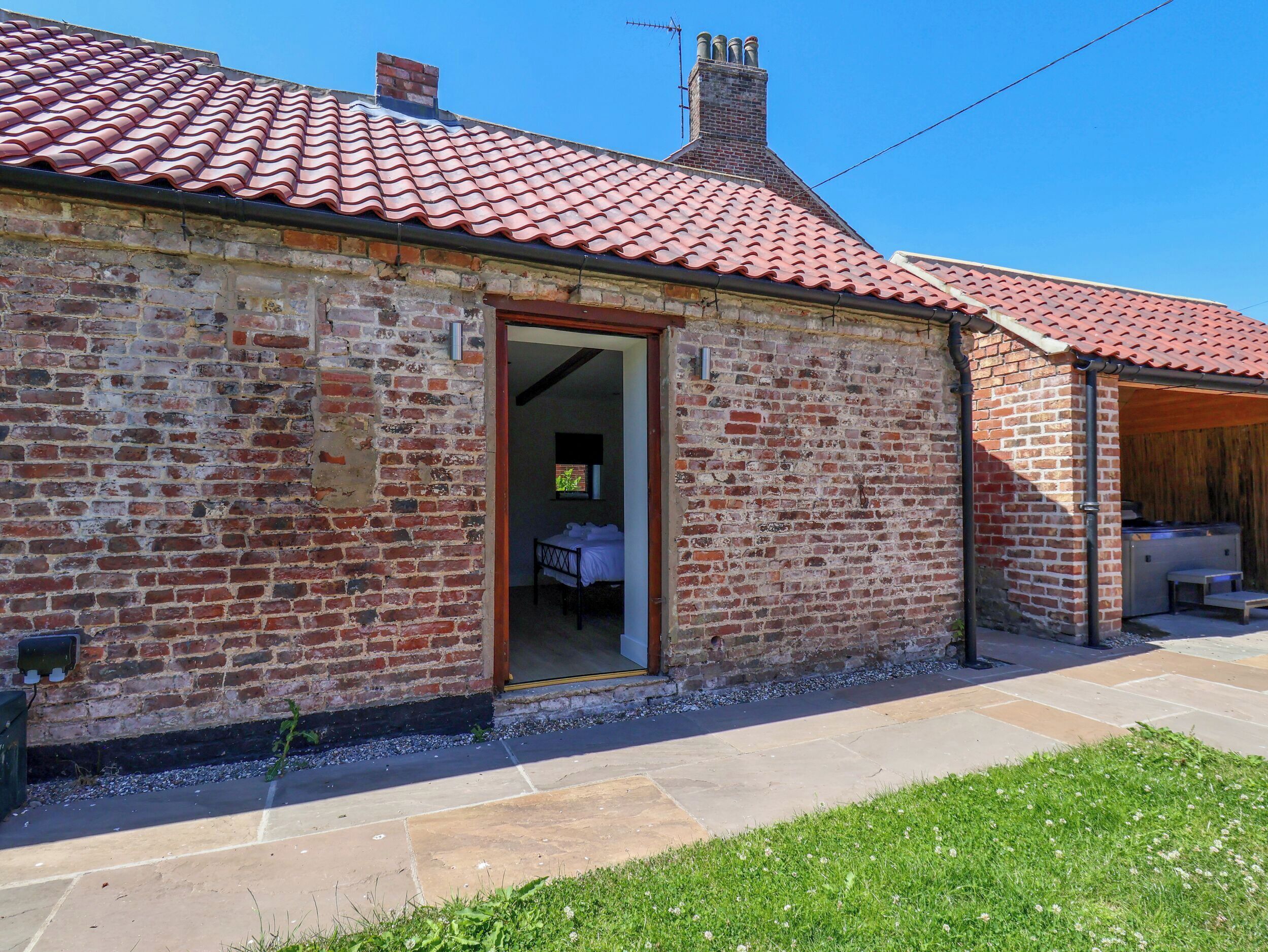 A brick building with a red-tiled roof, a visible bedroom with a bed, and a small outdoor seating area.