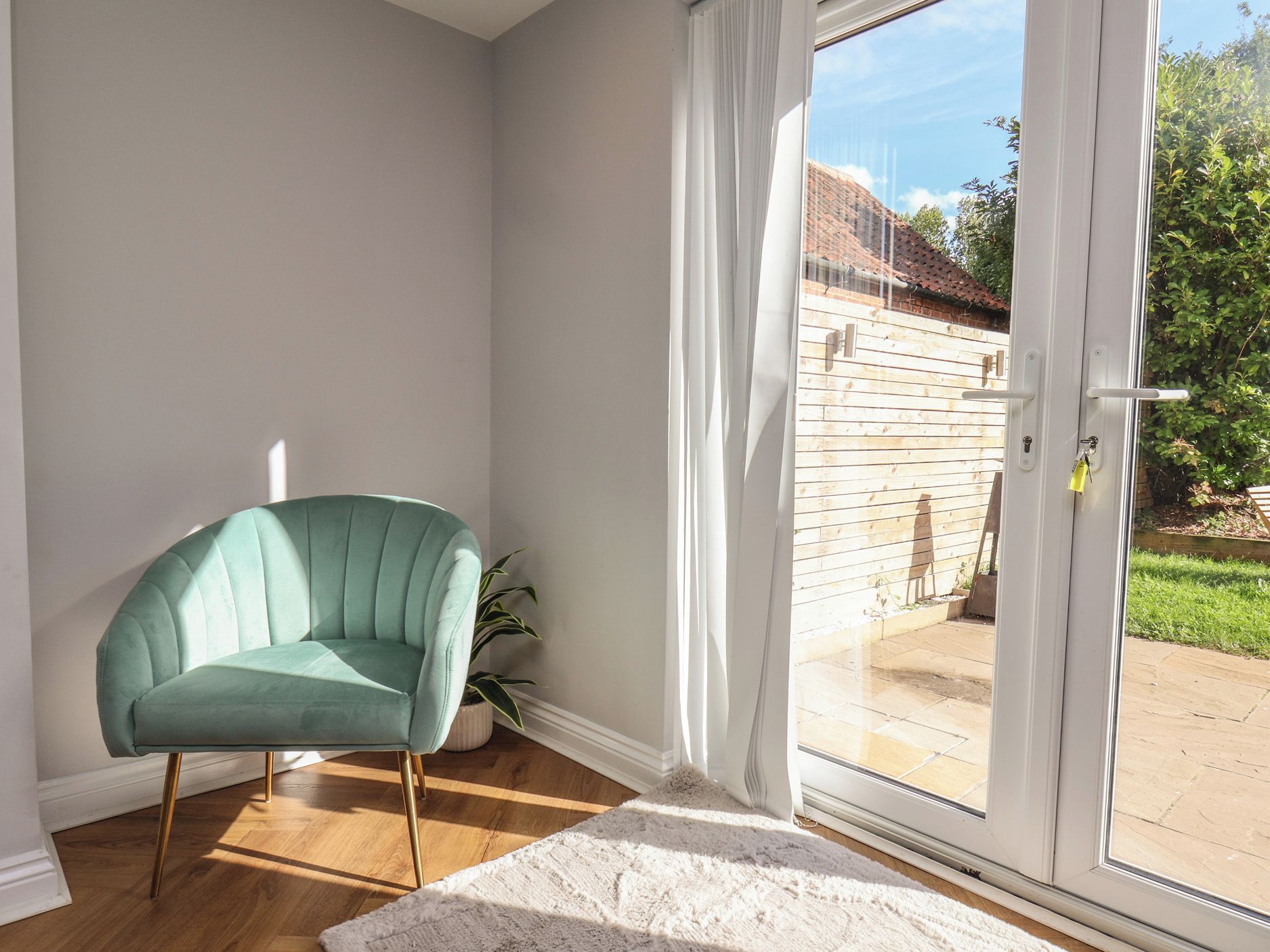 Una habitación con un sillón turquesa, una alfombra blanca y un ventanal corredero que da a un espacio exterior con pared de ladrillo y vegetación.