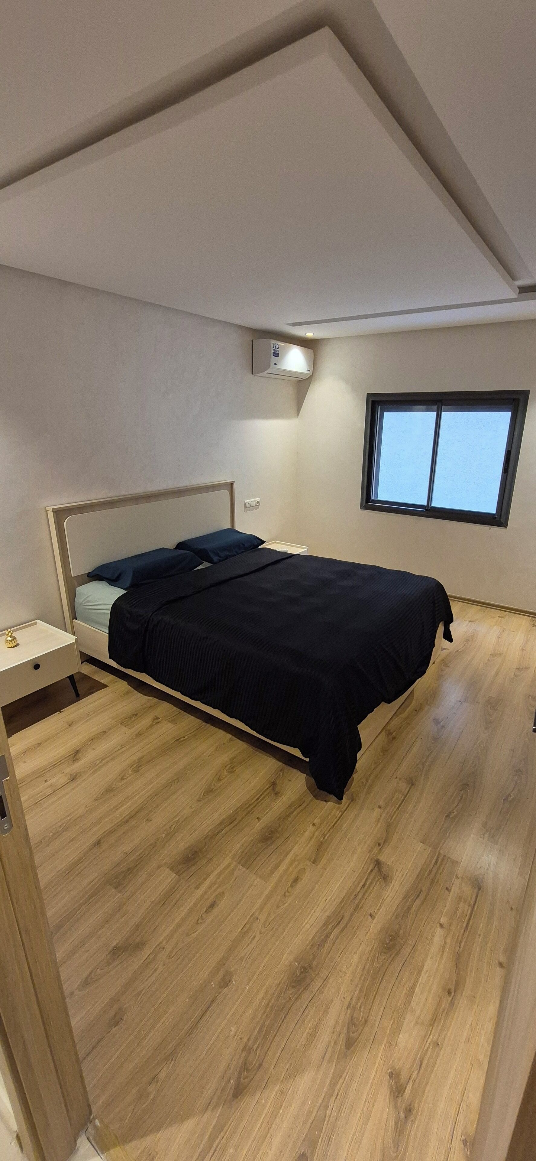 A bedroom with a bed, a bedside table, a window, and an air conditioning unit.