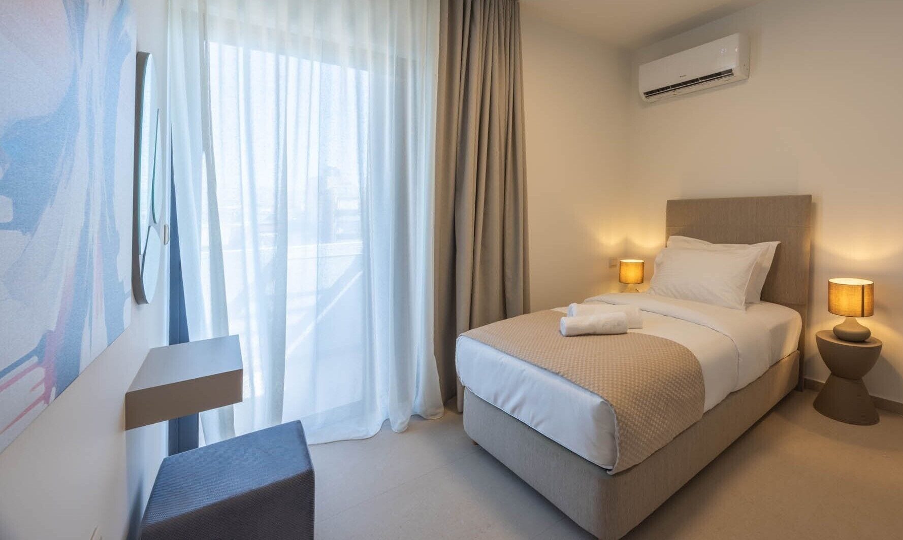 Habitación de hotel con cama, lámparas de noche, equipo de aire acondicionado y vistas a través de cortinas translúcidas.