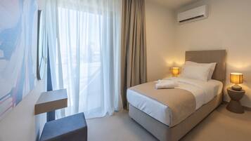 A hotel room with a bed, bedside lamps, an air conditioning unit, and a view through sheer curtains.